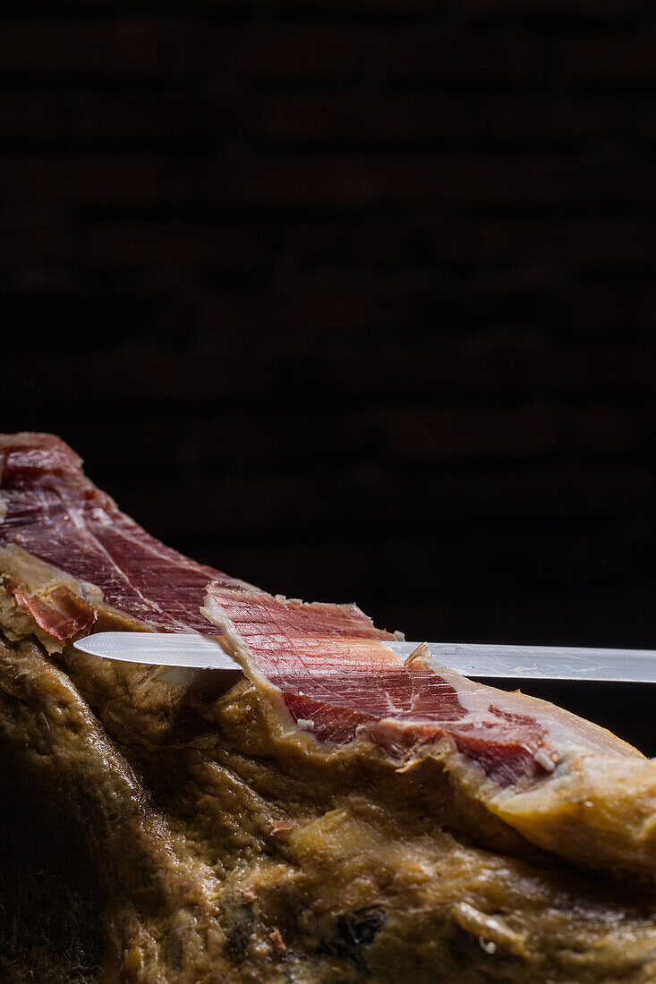 Crop unrecognizable cook cutting slice of tasty dry cured Spanish jamon on black background