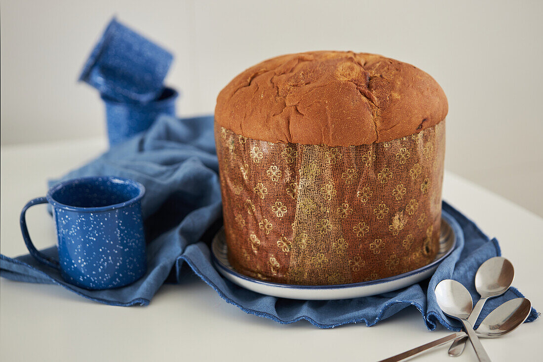 Ungeschnittener, frisch gebackener, handwerklich hergestellter Weihnachts-Panettone-Kuchen auf einem Teller, der auf einem blauen Tuch neben Tassen an einer weißen Wand steht