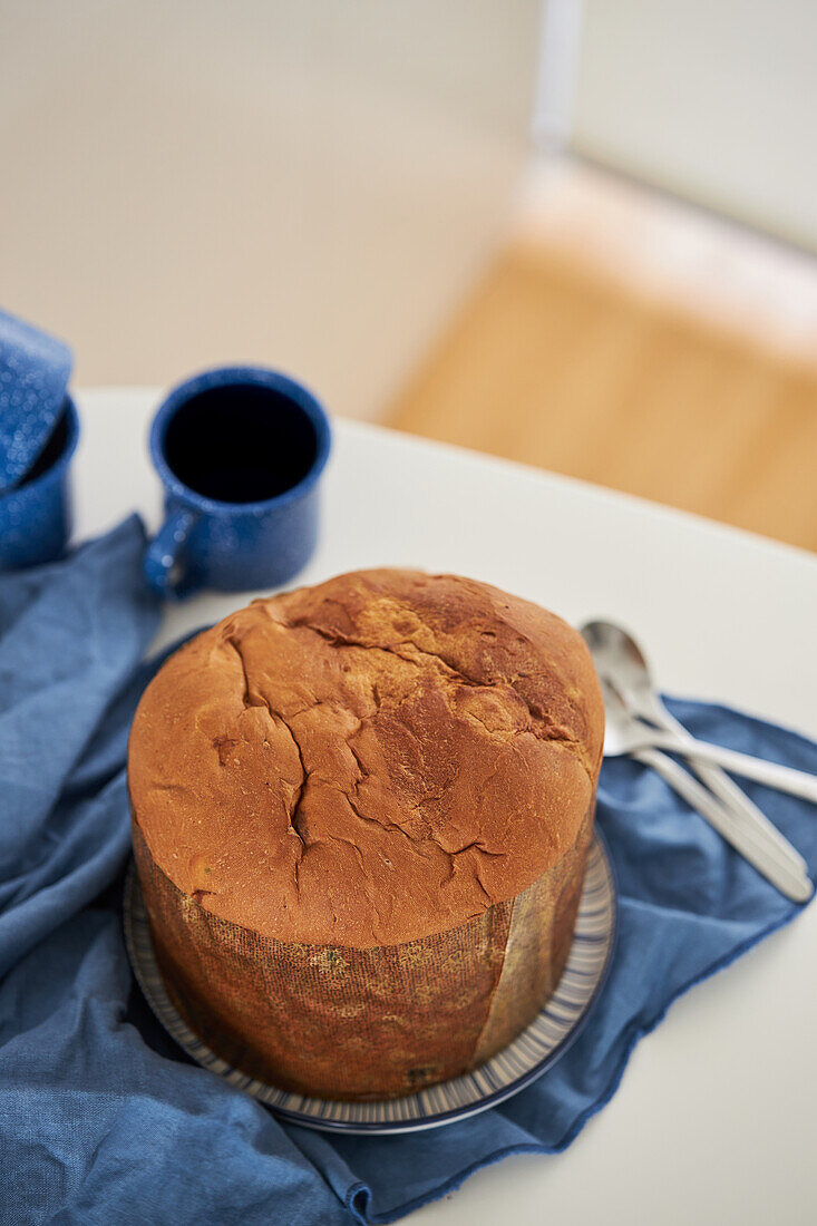 Blick von oben auf einen ungeschnittenen, frisch gebackenen, handwerklich hergestellten Weihnachts-Panettone-Kuchen auf einem Teller, der neben Tassen auf einem blauen Tuch liegt