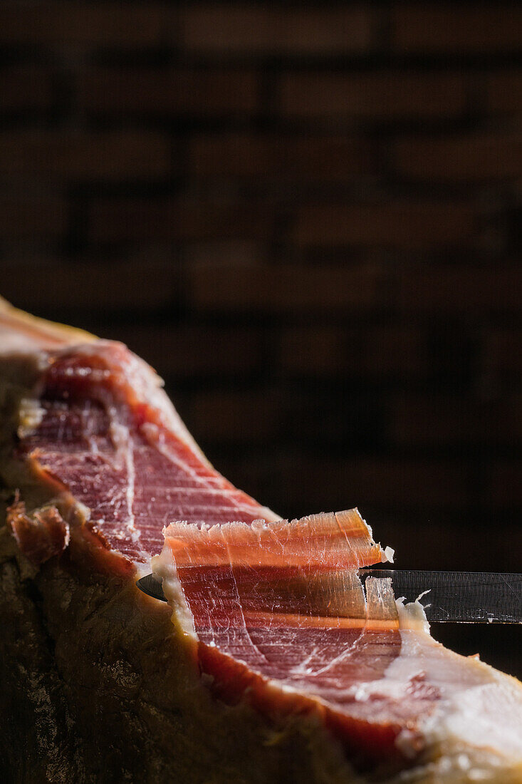 Crop unrecognizable cook cutting slice of tasty dry cured Spanish jamon on black background