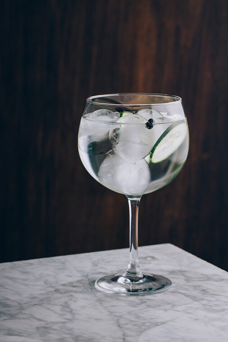 Glass of refreshing alcohol gin tonic cocktail with sliced cucumber and ice cubes on table