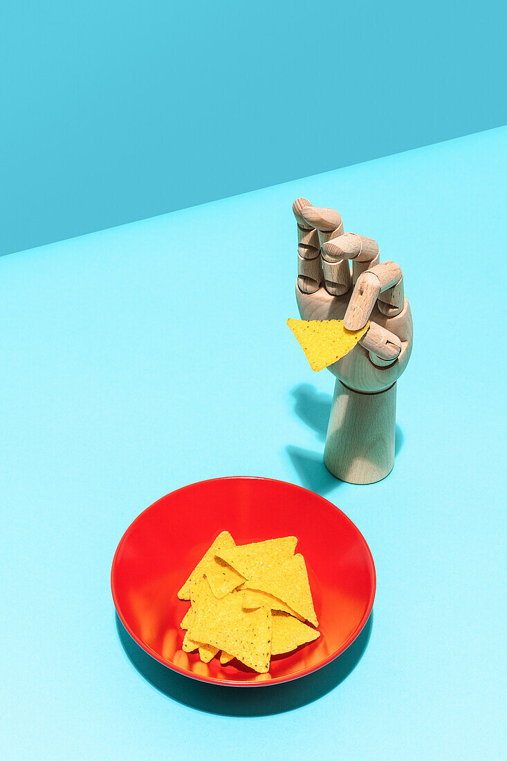 From above artificial hand holding tortilla chip placed near bowl with tortilla chips for Mexican cuisine dish on blue background