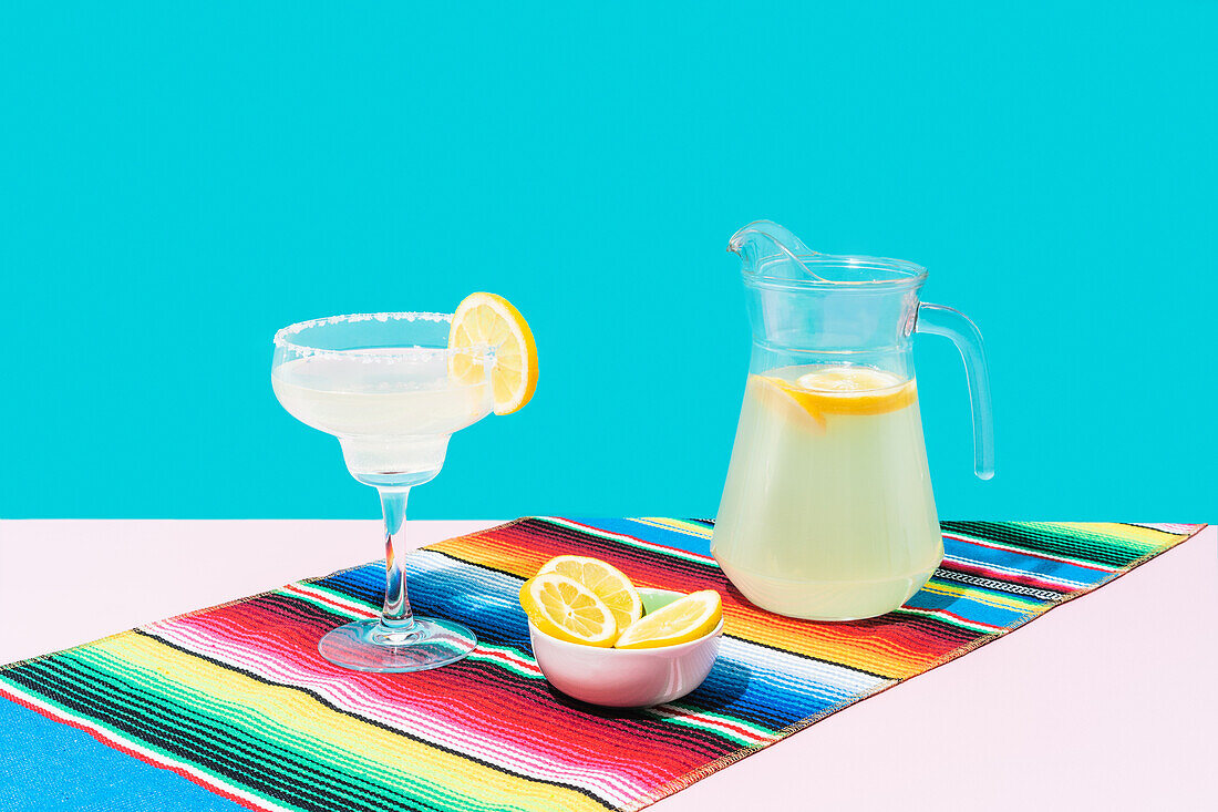 Glas und Krug mit kalter Limonade mit frischen Zitronen auf bunter mexikanischer Teppichdecke auf blauem Hintergrund