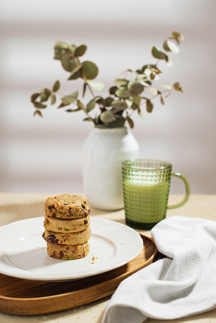 Teller mit einem Stapel leckerer Kekse und einem Becher frischer Milch auf dem Tisch neben Serviette und Vase mit Zweigen am Morgen