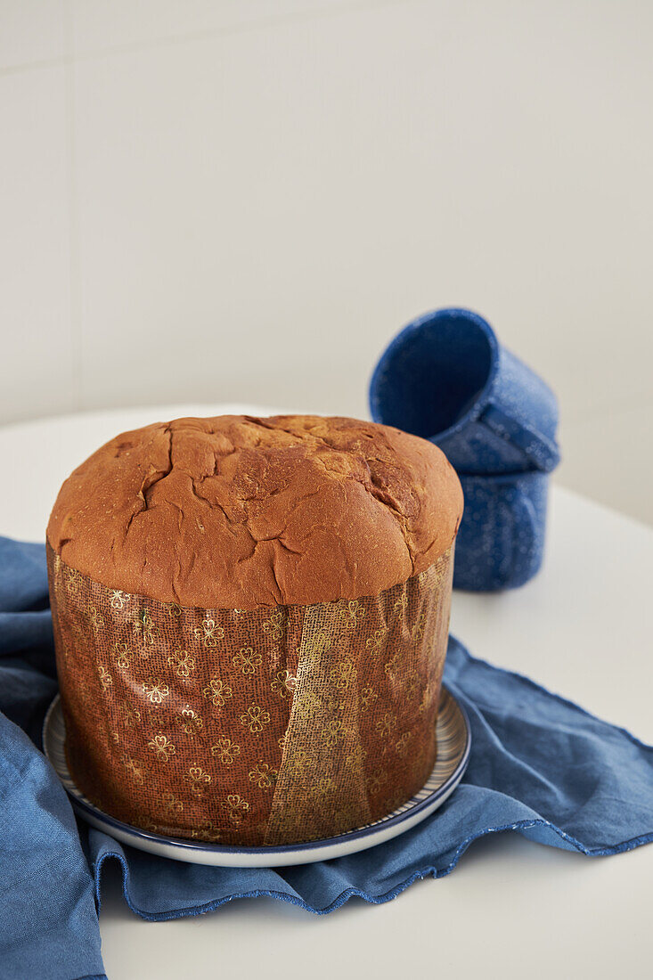 High angle of uncut fresh baked artisan Christmas panettone cake on plate placed on blue cloth next to cups against white wall