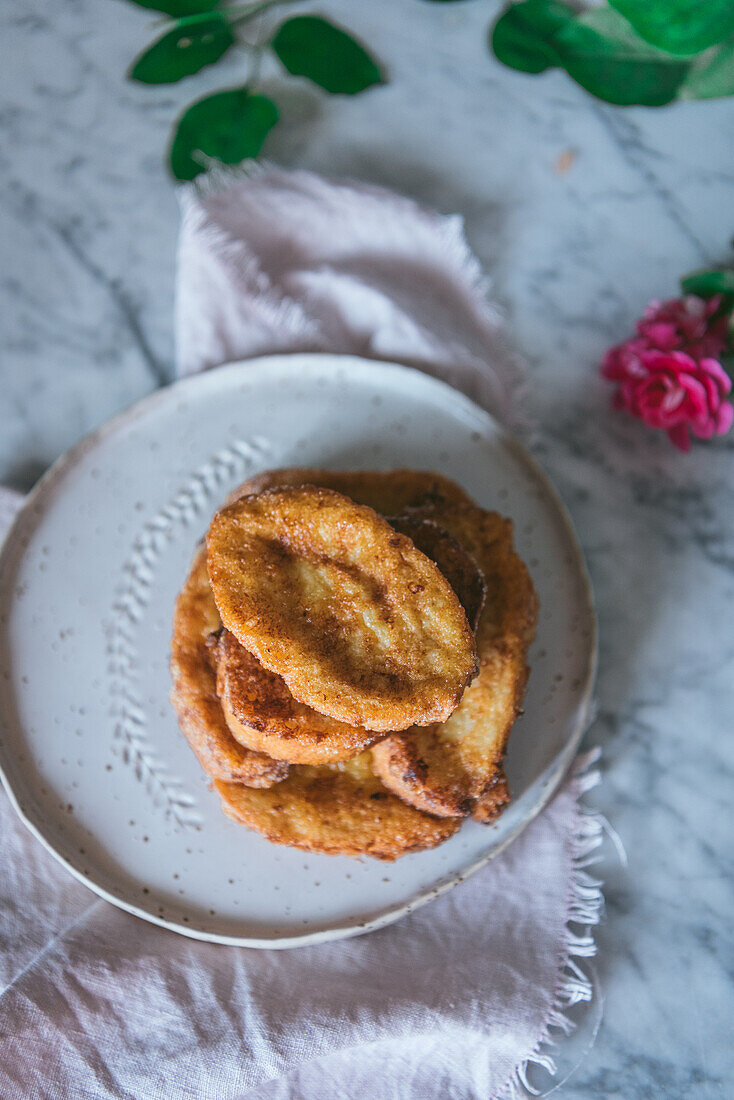 Draufsicht auf ein hausgemachtes traditionelles spanisches Osterdessert mit französischem Toast, serviert auf einem Teller neben einer Rosenblüte auf marmoriertem Hintergrund