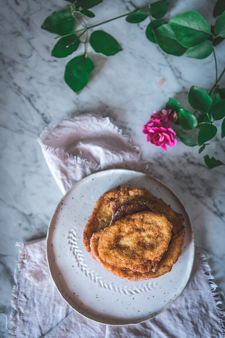 Draufsicht auf ein hausgemachtes traditionelles spanisches Osterdessert mit französischem Toast, serviert auf einem Teller neben einer Rosenblüte auf marmoriertem Hintergrund