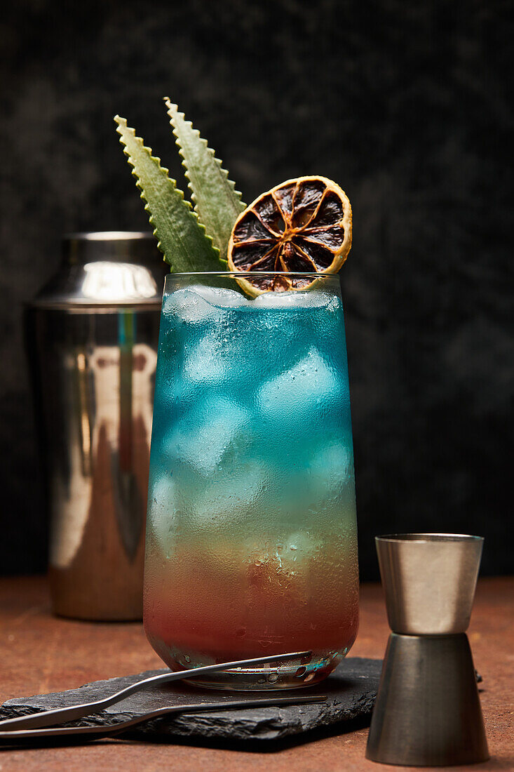 Glas Regenbogenparadies mit buntem Cocktail, garniert mit einer Orangenscheibe neben einem Jigger auf einem Metalltablett auf dem Tisch
