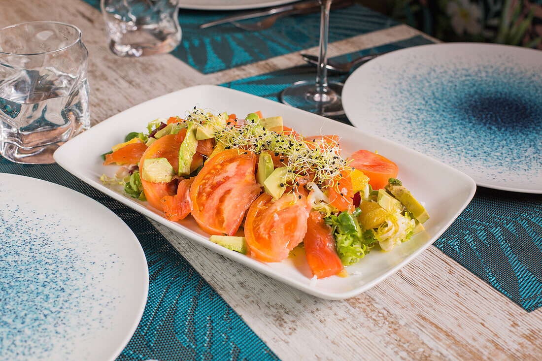 Delicious salad with sliced fresh tomatoes and avocado with marinated salmon under sprouts near glass of red wine on table