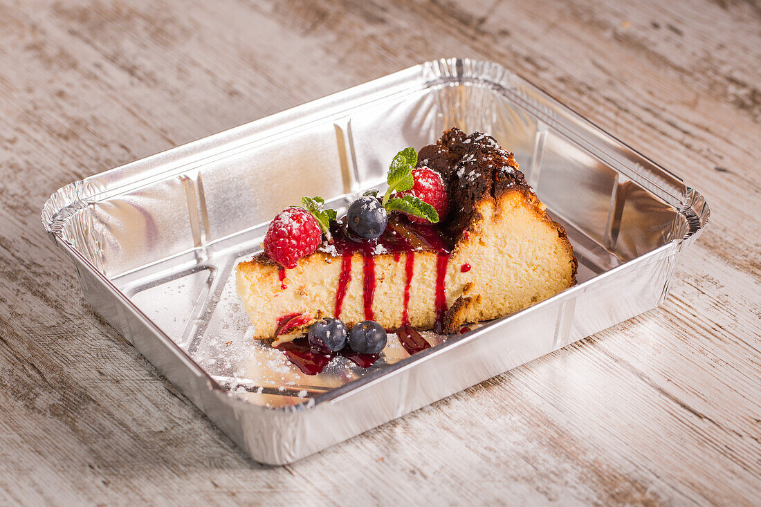 Tasty cheesecake with red jam and ripe raspberries with blueberries near fresh mint leaves and powdered sugar in aluminum baking pan