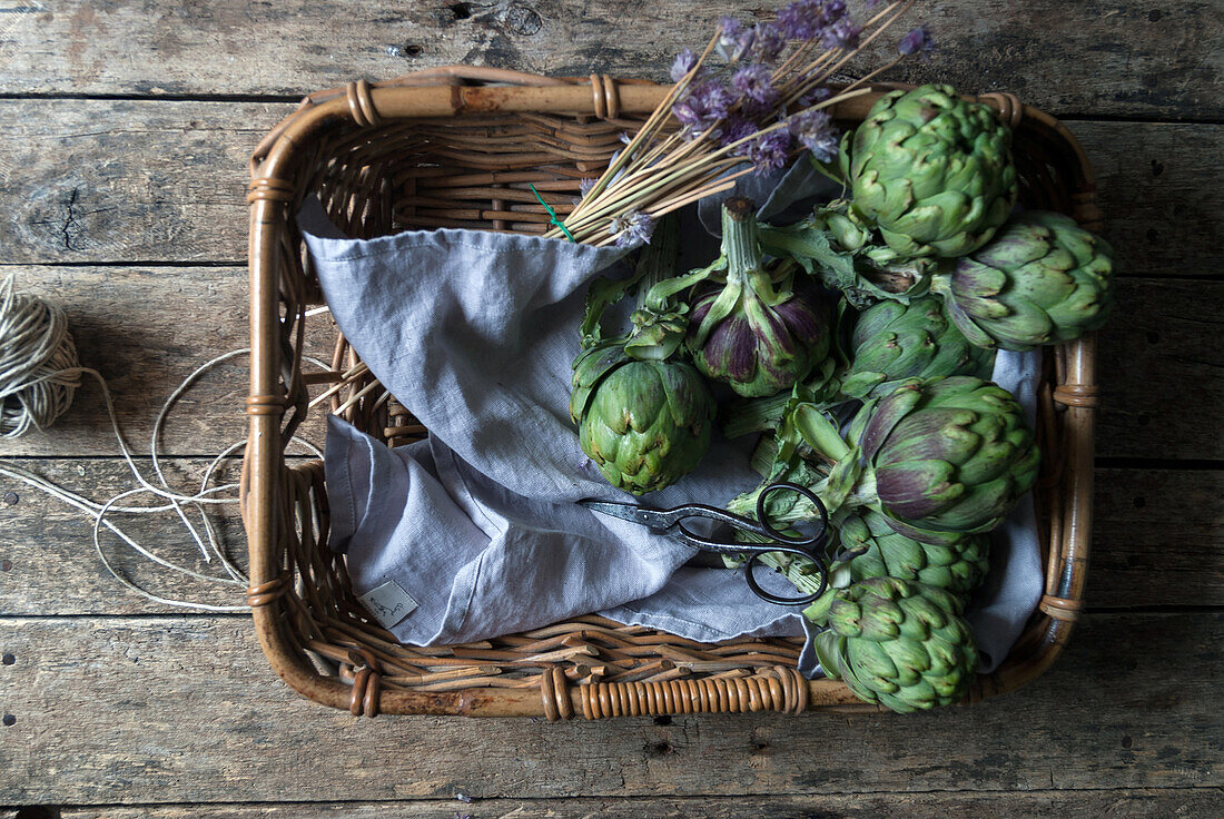 From above view of fresh green artichokes laid in wicker basket on napkin decorated with flowers on wooden background