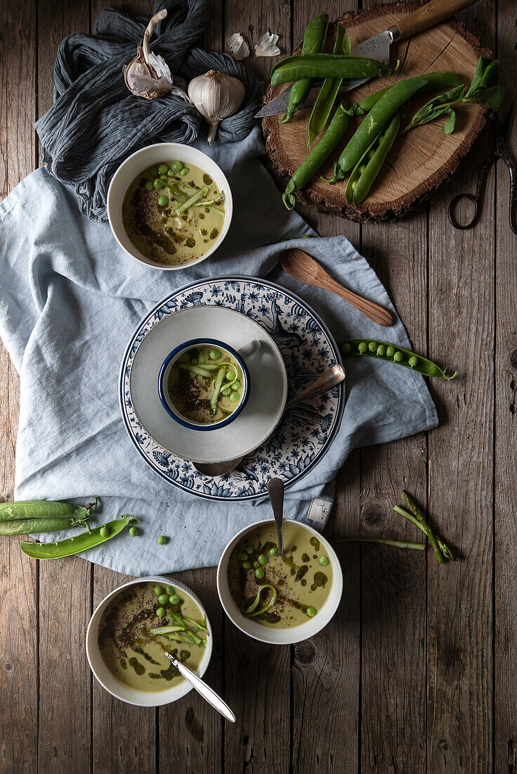 Flat lay of bowls with green pea and coconut cream soup on wooden table with pea pods and garlic in composition