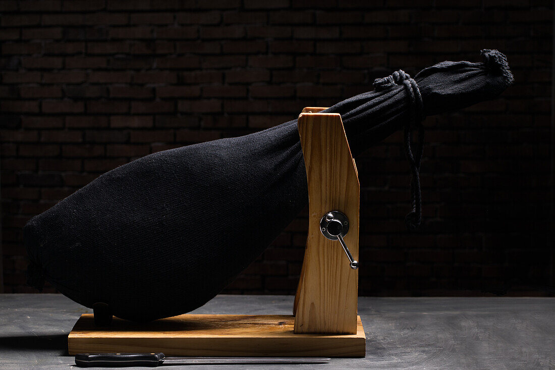 Delicious dry cured Spanish pork leg wrapped in black bag on wooden holder on gray surface