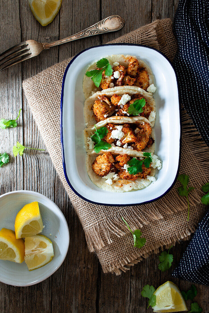 From above cauliflower snack tacos in bowl and lemon pieces in plate on wooden table