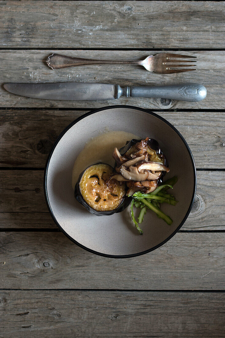 Top view of served round bowl with eggplant and shiitake mushrooms on wooden table with silver cutlery