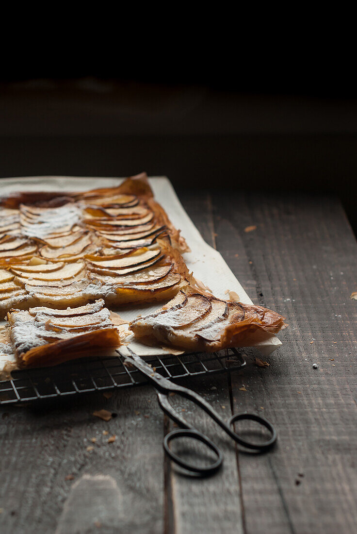 Nahaufnahme eines Kühlregals mit Frangipane-Apfelkuchen auf Pergament, der mit einer alten Schere auf einem Holztisch liegt