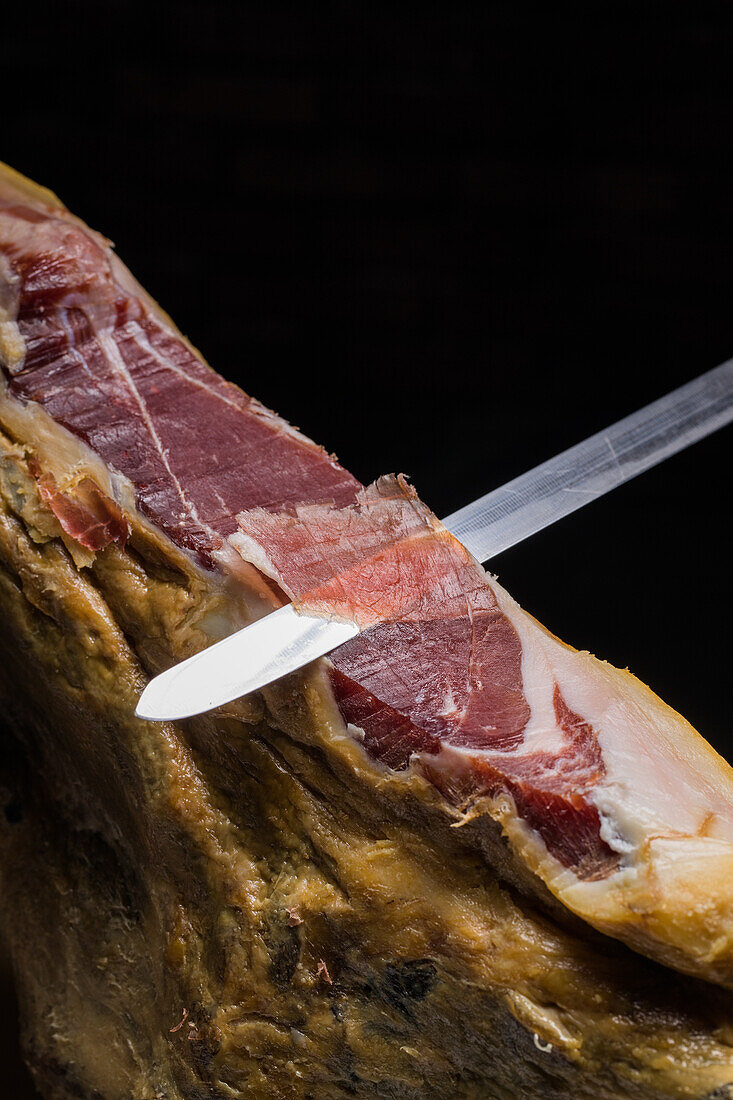 Crop unrecognizable cook cutting slice of tasty dry cured Spanish jamon on black background