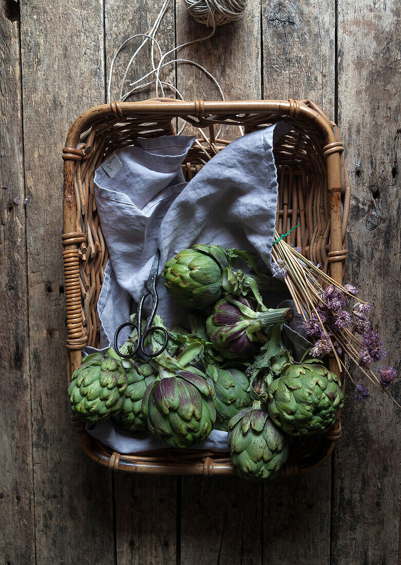 From above view of fresh green artichokes laid in wicker basket on napkin decorated with flowers on wooden background