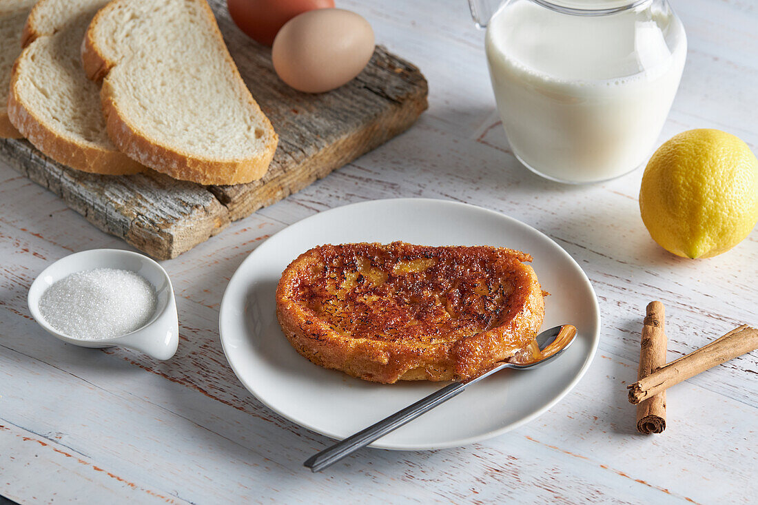 Blick von oben auf einen Teller mit appetitlichem spanischem Torrija-Brot, das mit rohen Eiern und Zucker auf dem Tisch liegt, neben einem hölzernen Schneidebrett mit einem Krug frischer Milch und Brotscheiben