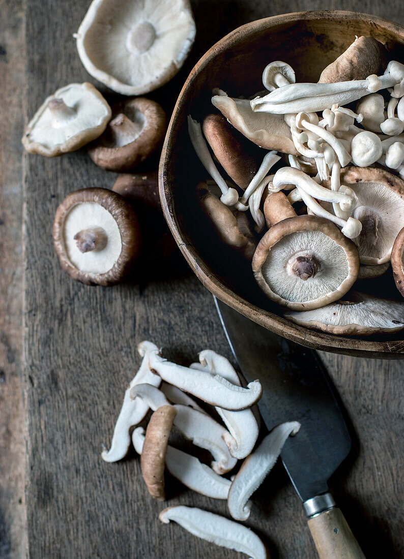 Draufsicht auf eine Holzschale voller Shiitake-Pilze auf einem Holztisch mit einem Messer darauf