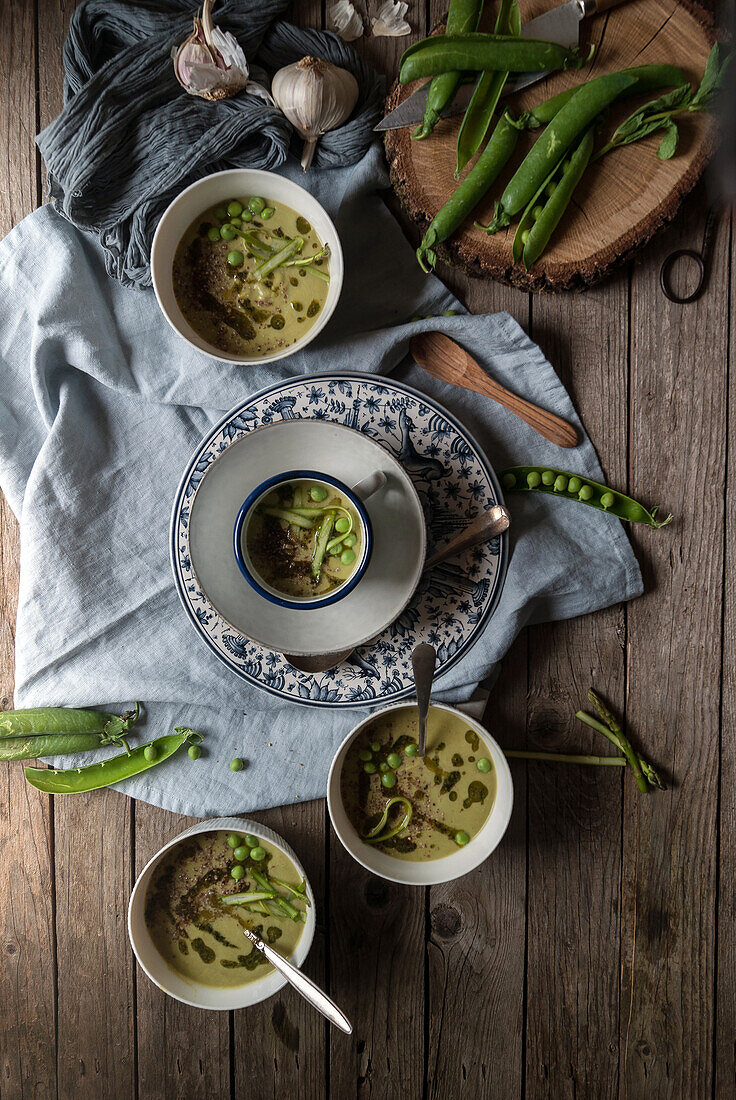 Flache Schalen mit grüner Erbsen- und Kokosnusscremesuppe auf Holztisch mit Erbsenschoten und Knoblauch in Komposition