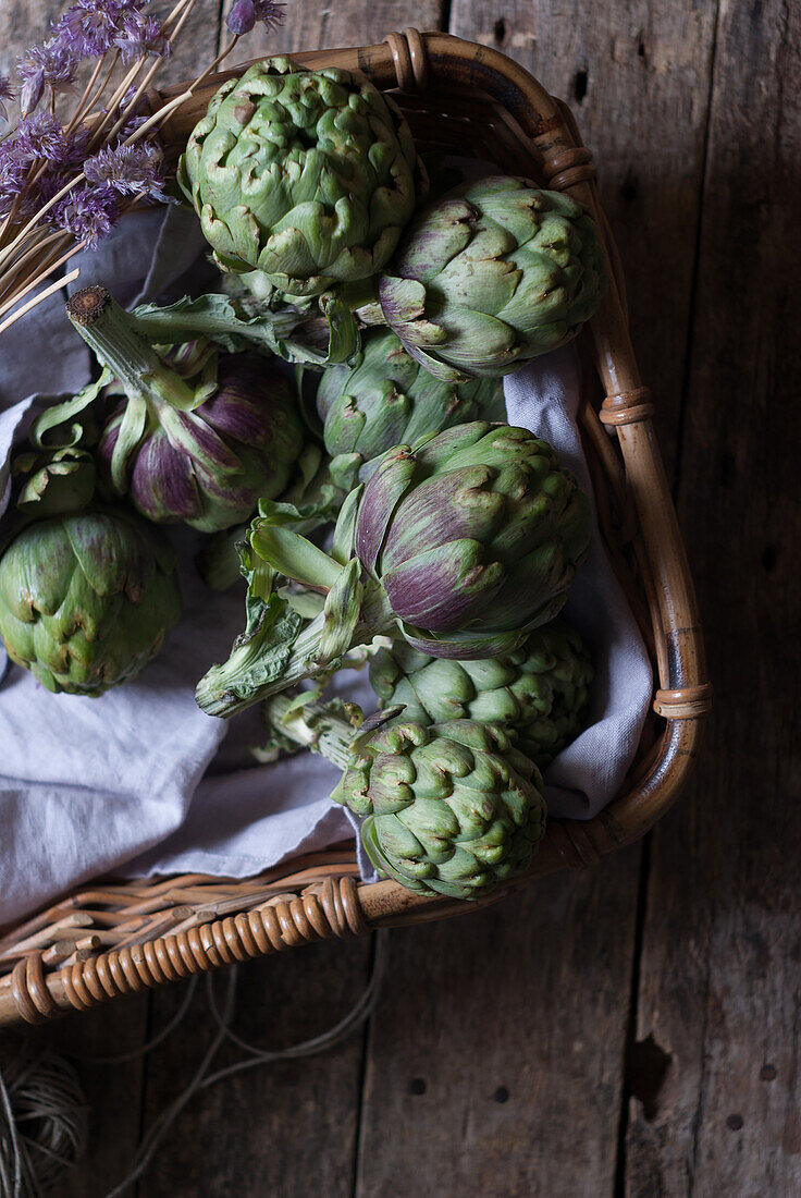 Closeup from above view of green artichokes laid in wicker basket with bunch of little purple flowers