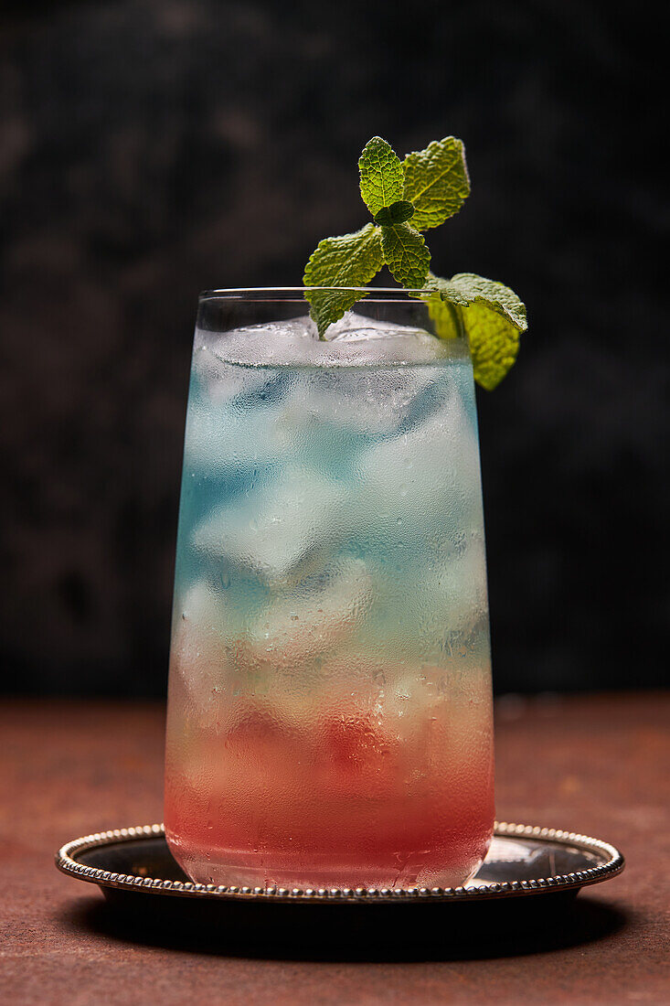 Glass of rainbow paradise colorful cocktail garnished with mint leaf on metal tray