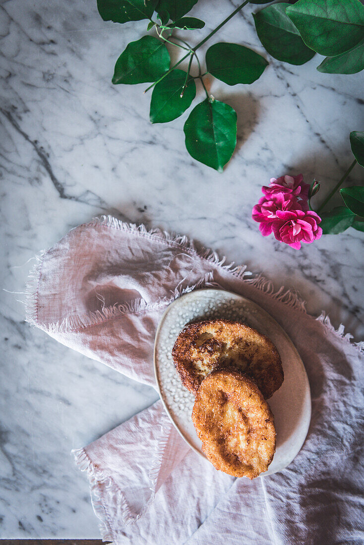 Draufsicht auf ein hausgemachtes traditionelles spanisches Osterdessert mit französischem Toast, serviert auf einem Teller neben einer Rosenblüte auf marmoriertem Hintergrund