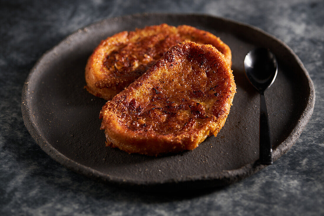 Hoher Blick auf köstliche hausgemachte Torrija mit knuspriger Kruste auf schwarzem Teller mit Löffel auf Marmortisch