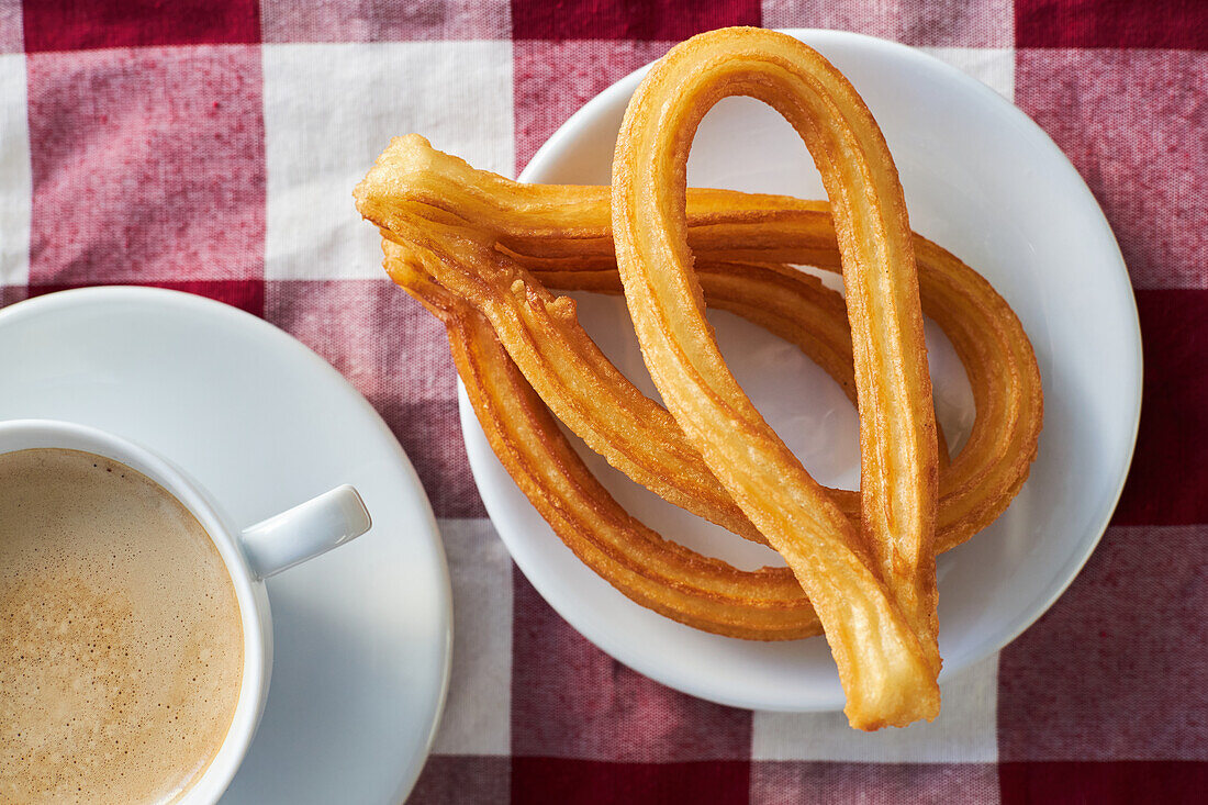 Appetitliche Churros auf einem Teller und eine Tasse frischen Morgenkaffee auf einer Serviette auf dem Frühstückstisch in der Küche