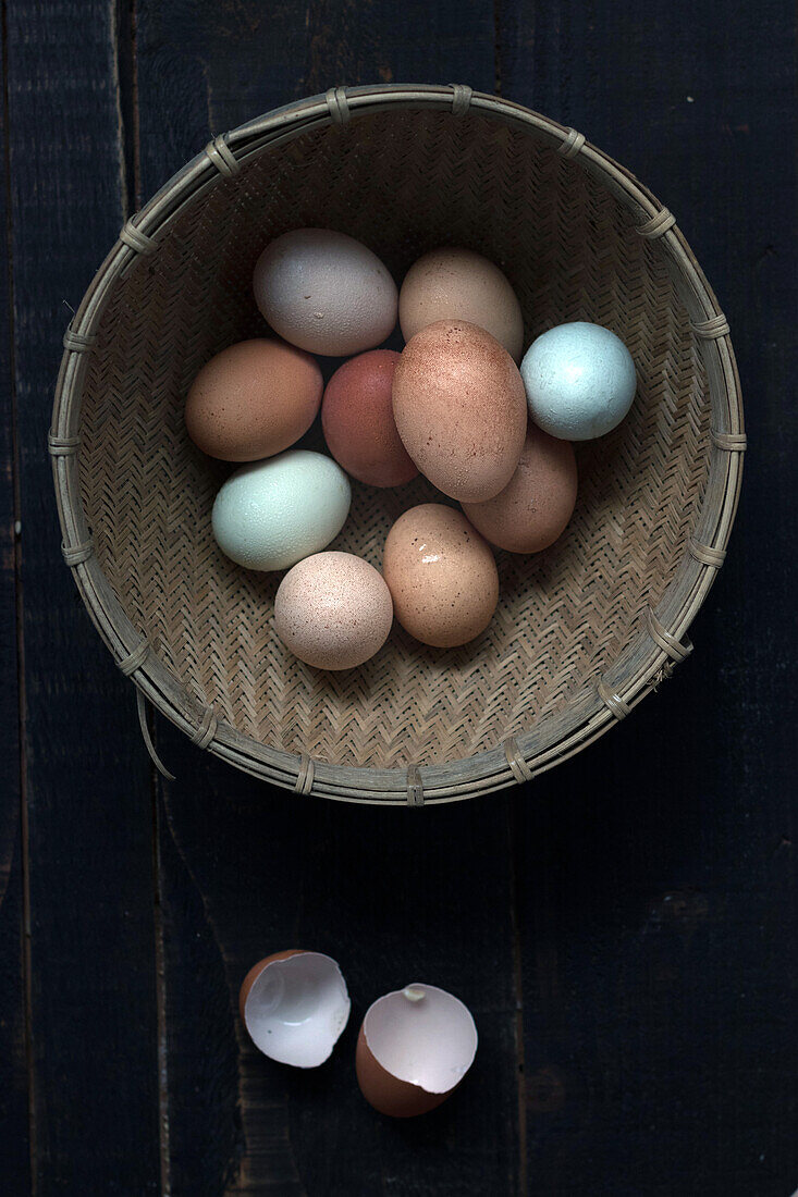 From above shot of wicker bowl filled with raw eggs on wooden table with eggshell near