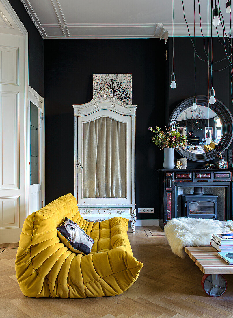 Yellow designer sofa and black walls in living room with antique cupboard and fireplace