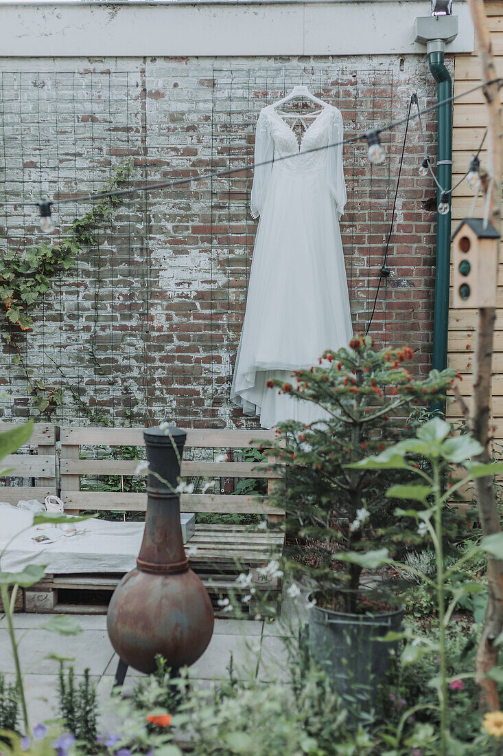 Bride's dress hanging on trellis in garden with rusty vase and plants in foreground