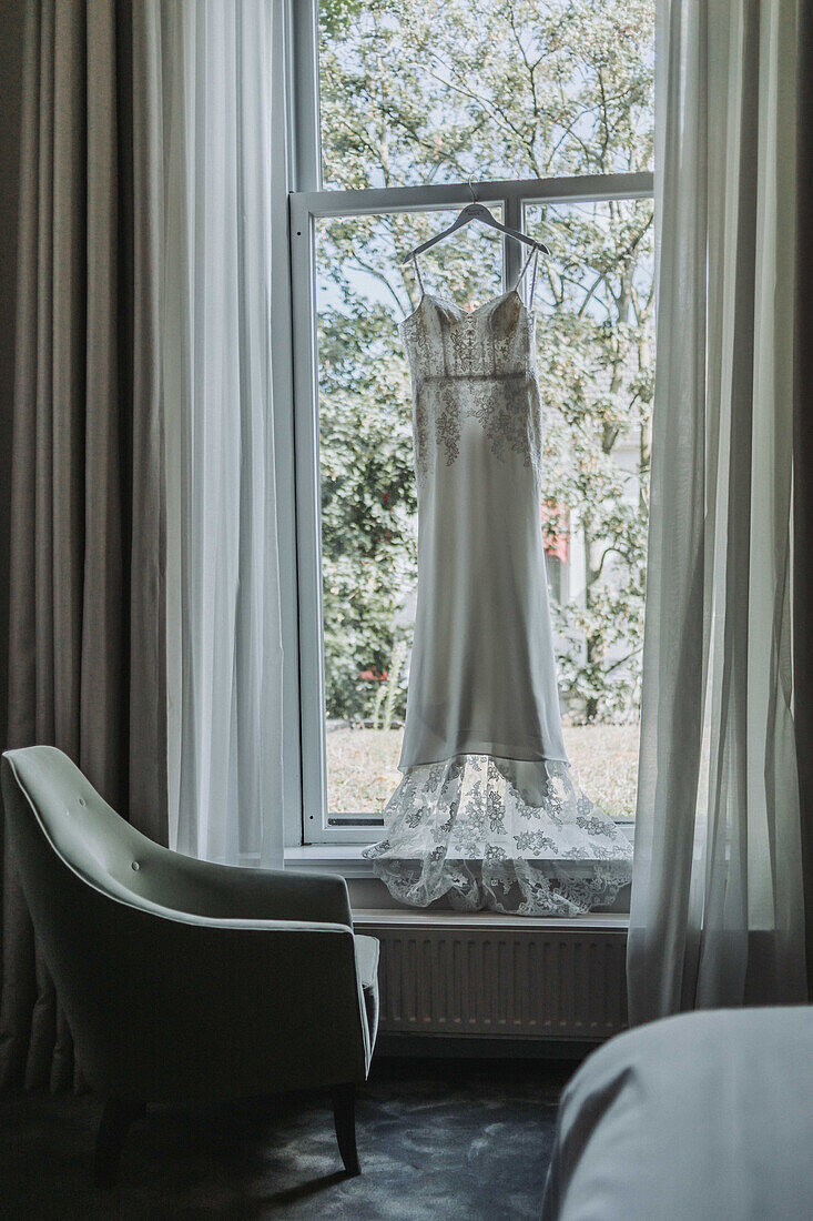 Wedding dress hangs on window in room with green armchair and curtains