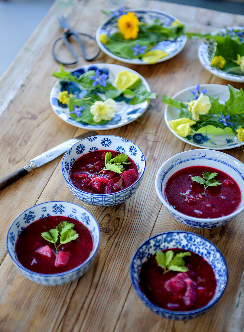 Holztisch mit Rote-Bete-Suppe in blauen Schalen und Tellern mit essbaren Blüten