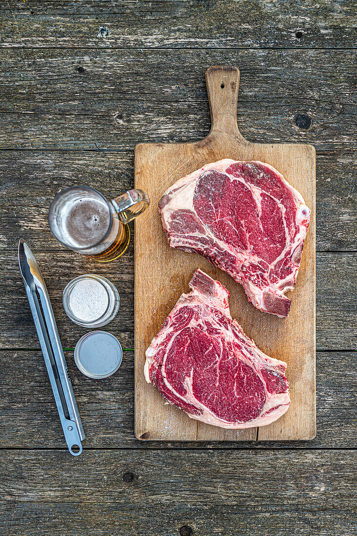 Ochsenkoteletts zum Grillen daneben Bier und Grillzange