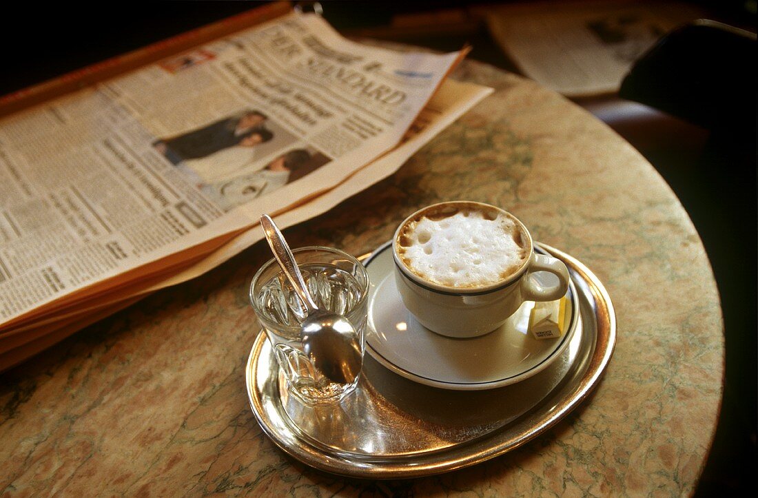 Tasse Cappuccino, Glas Wasser & Zeitung auf Kaffeehaustisch