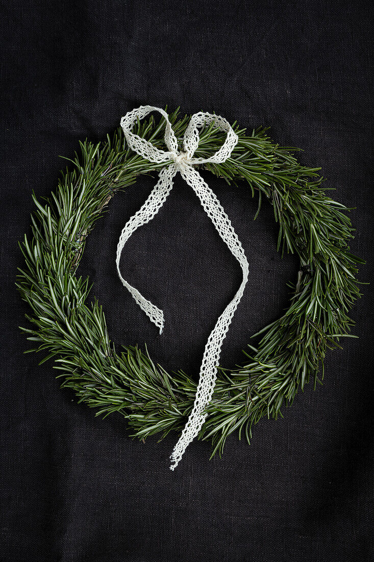 Gebundener Rosmarinkranz (Rosmarinus officinalis) mit weißer Spitzenschleife vor schwarzem Hintergrund