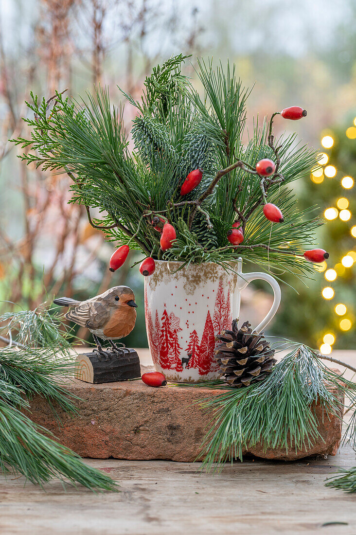 Strauß aus Hagebutten, Seidenkiefernzweigen (Pinus Strobus) in Kaffeetasse und Vogelfigur als Winterdeko auf der Terrasse