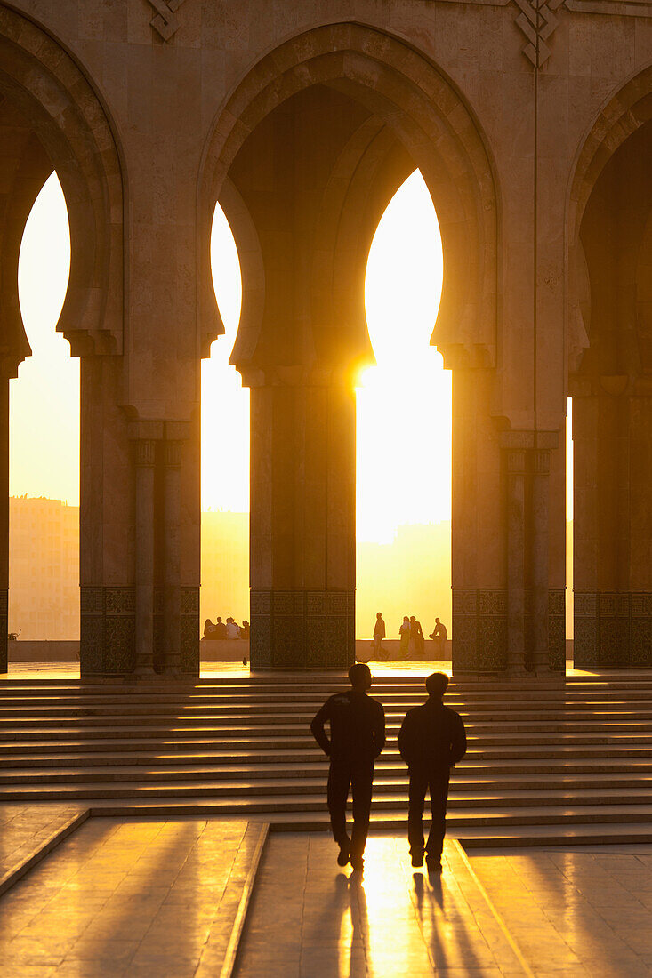 Silhouette of two people walking towards steps and archways with the sun glowing; Casablanca, Morocco