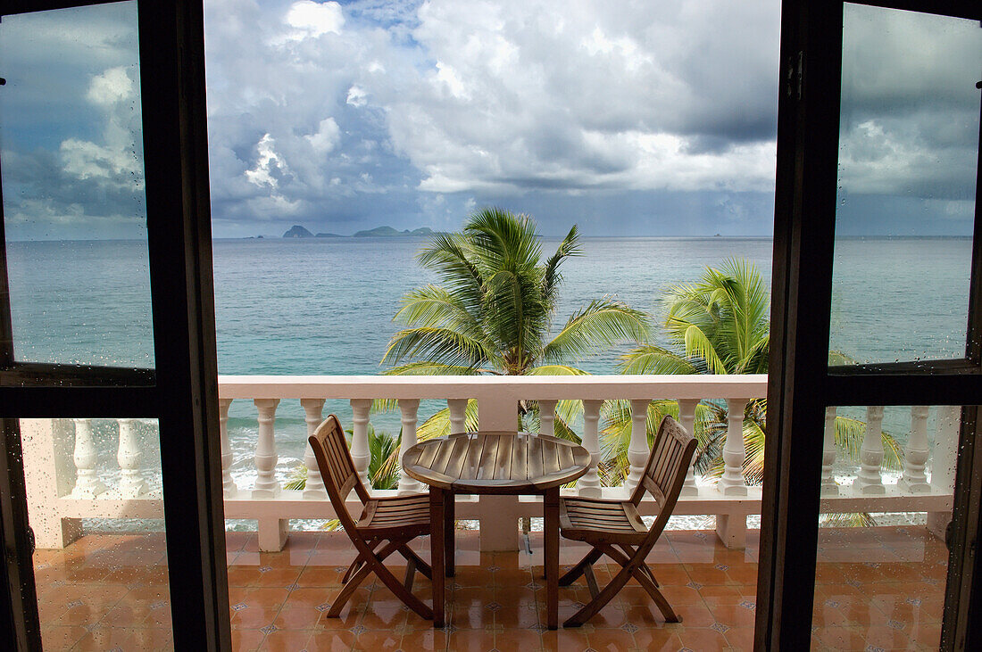 Blick durch eine Türöffnung auf eine Terrasse mit Tisch und Stühlen im Petite Anse Hotel. Sturmwolken sammeln sich über dem Ozean; Grenada, Karibik