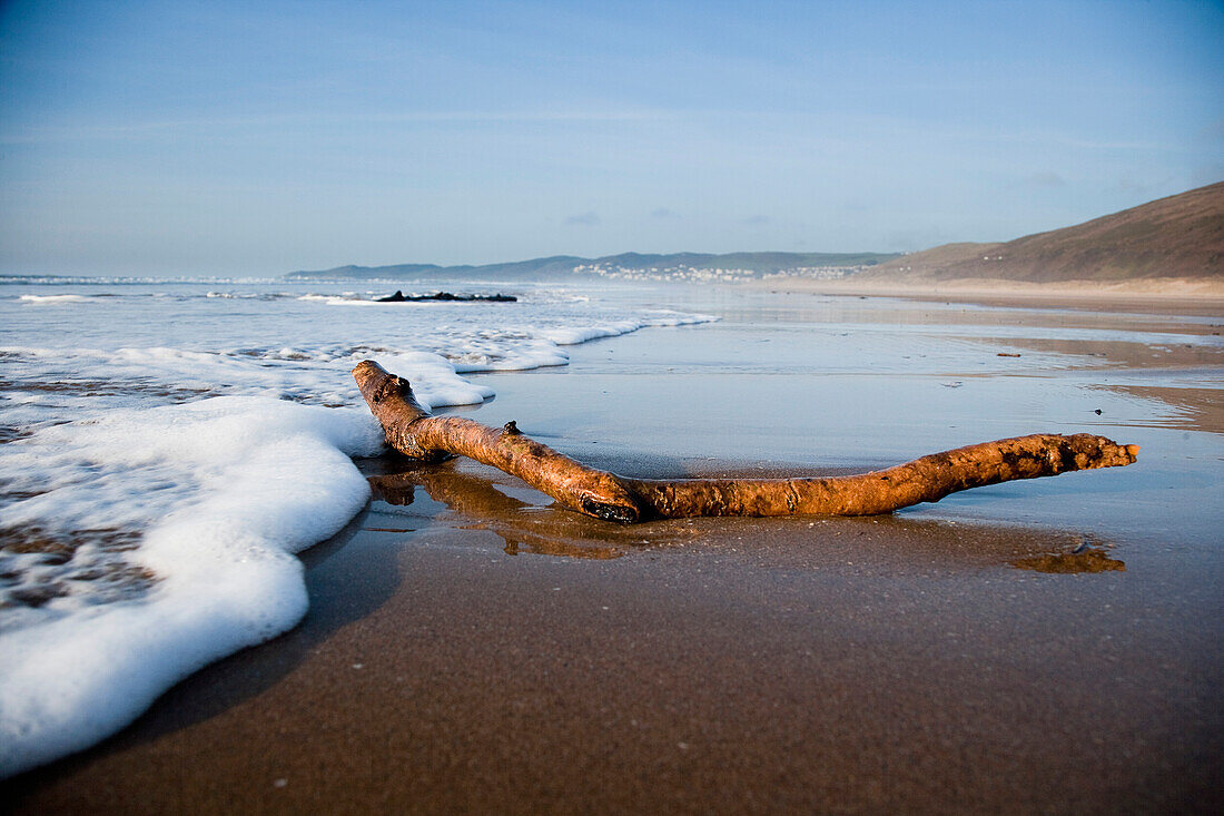 Wellen plätschern am Sandstrand gegen Treibholz in Putsborough Sands, Nord-Devon, Großbritannien