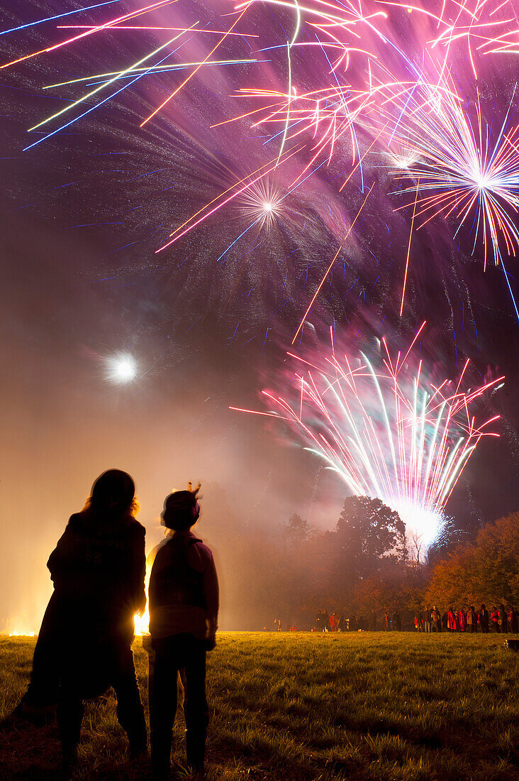 Silhouetten von zwei Menschen vor einem großen Lagerfeuer und Feuerwerk bei der Ewhurst and Staplecross Bonfire Night, East Sussex, Großbritannien