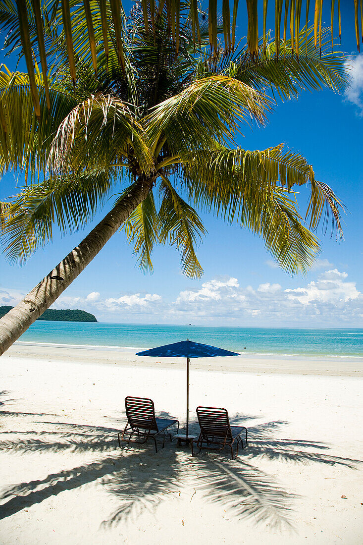 Deck Chairs Under Parasol On White Sandy Beach With Palm Trees Overlooking Blue Sea. Pantai Cenang (Cenang Beach), Pulau Langkawi, &#10; Malaysia, South East Asia.