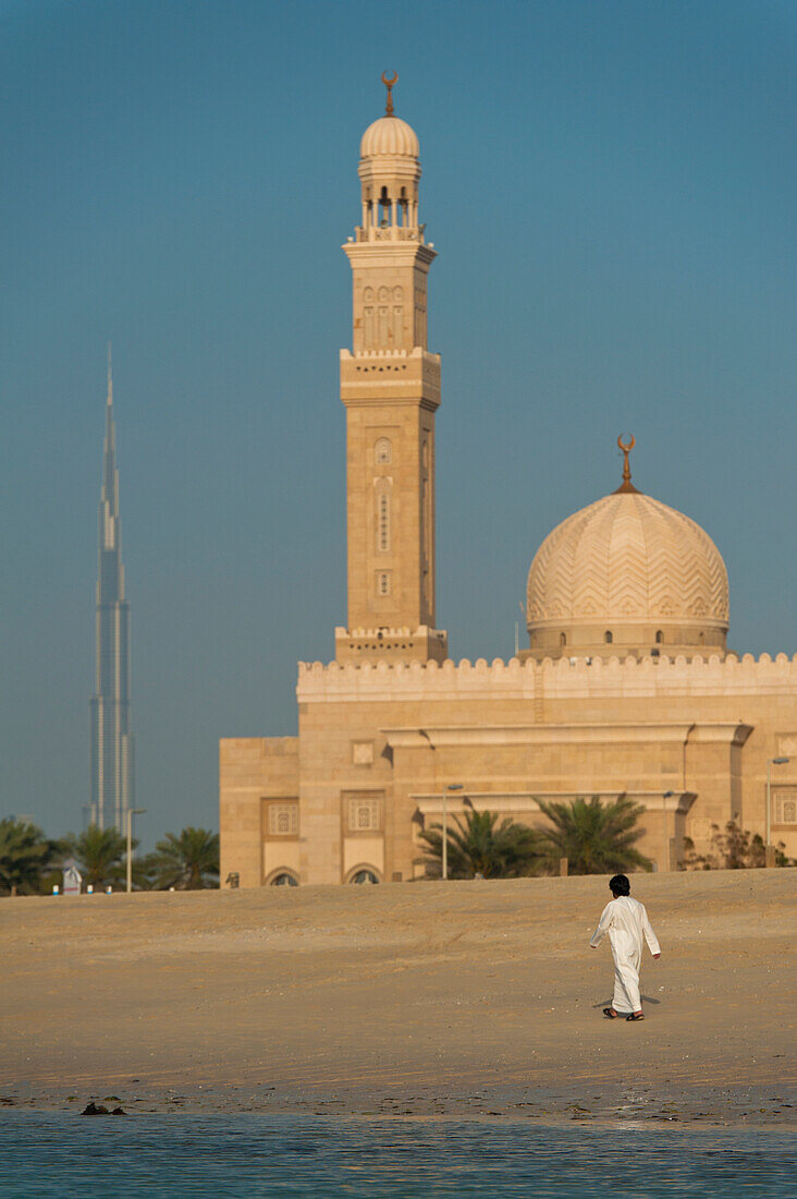 Dubai, Uaeman Walking Along Beach In Front Of Small Mosque And The Burj Khalifa In The Distance