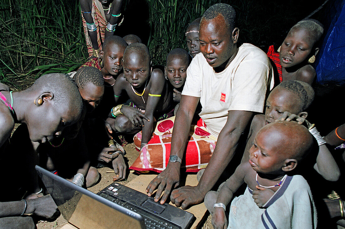 Group of Mursi tribal children looking in owe at the first Mursi owned laptop computer been operated in the deep of night. Makki / South Omo / Southern Nations, Nationalities & People's Region (Ethiopia).
