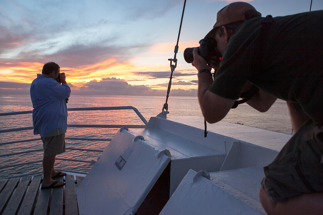 Two passengers on a tourist expedition vessel photographing sunset over the Pacific Ocean.; Pacific Ocean, Galapagos Islands, Ecuador