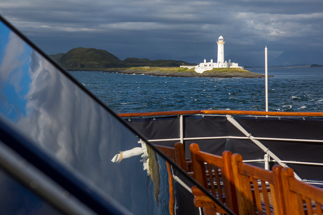 A lighthouse off the coast of Isle of Mull, Scotland, is illuminated against a dark sky, viewed from the deck of a ship; Isle of Mull, Scotland