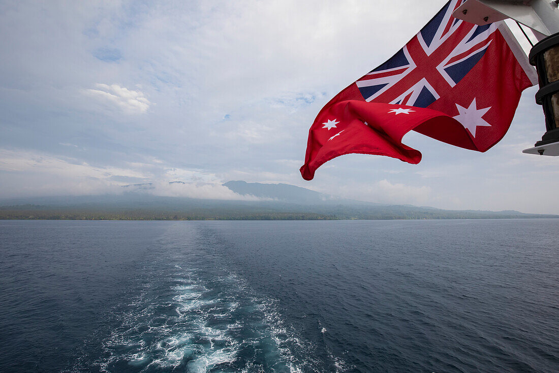 Rote australische Flagge am Heck eines Expeditionsschiffs, das um die Insel Karkar in der Bismarcksee vor der Nordküste Papua-Neuguineas segelt; Insel Karkar, Madang, Papua-Neuguinea