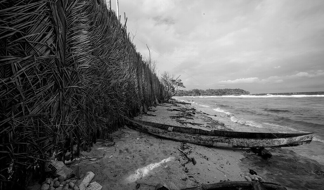 A beached canoe and a wall of woven sago palms (Cycas revoluta) built as a windbreak to protect a village on the windward coast of Tuam Island one of the Siassi Islands in the Bismarck Sea off the north coast of Papua New Guinea; Siassi, Vitiaz Strait, Papua New Guinea